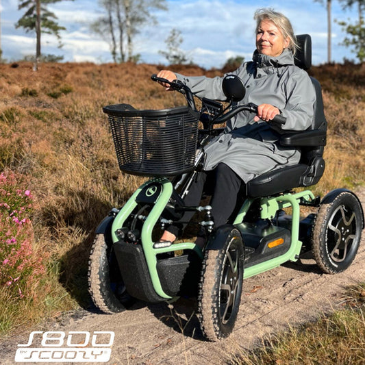 A woman wearing a gray raincoat sits on a STERLING S800 by Scoozy All-Terrain Mobility Scooter with a front basket, riding outdoors on a dirt path surrounded by heather and trees.