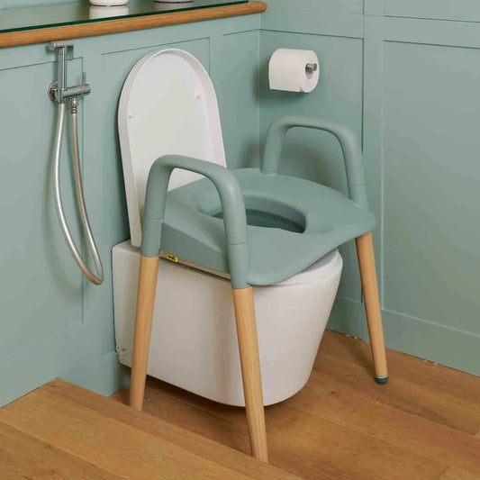 Spryly Poise raised toilet seat with armrests and wooden legs in sage over a toilet in a bathroom setting