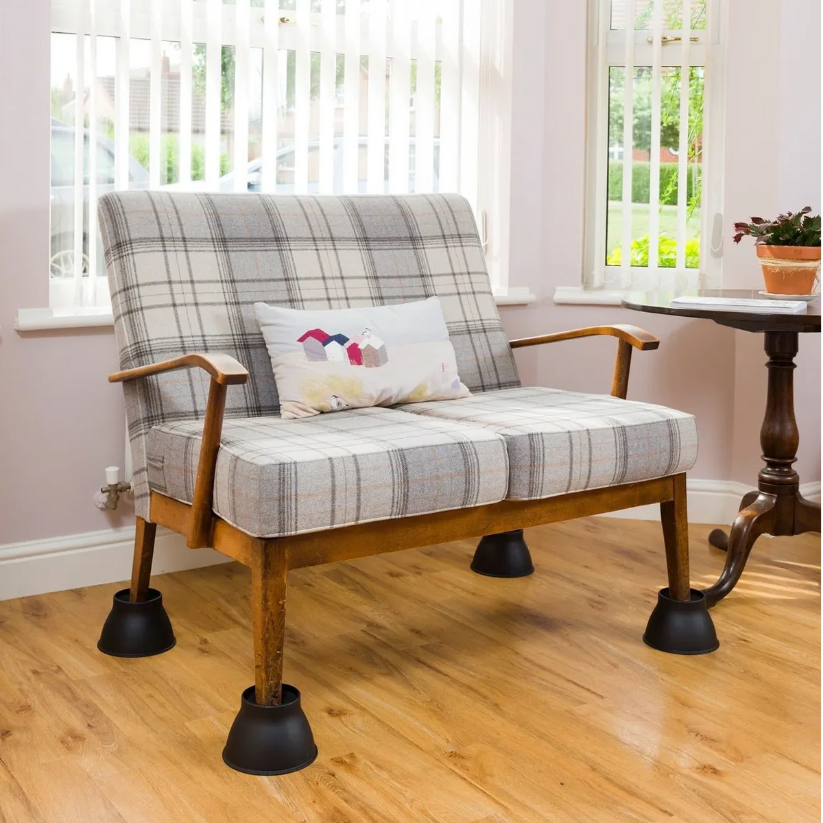 A wooden armchair with a gray and white plaid upholstered seat and back on Derby Elephant Feet Furniture Raiser 9cm, shown in a bright room with a light wood floor, white blinds, and a small decorative pillow.