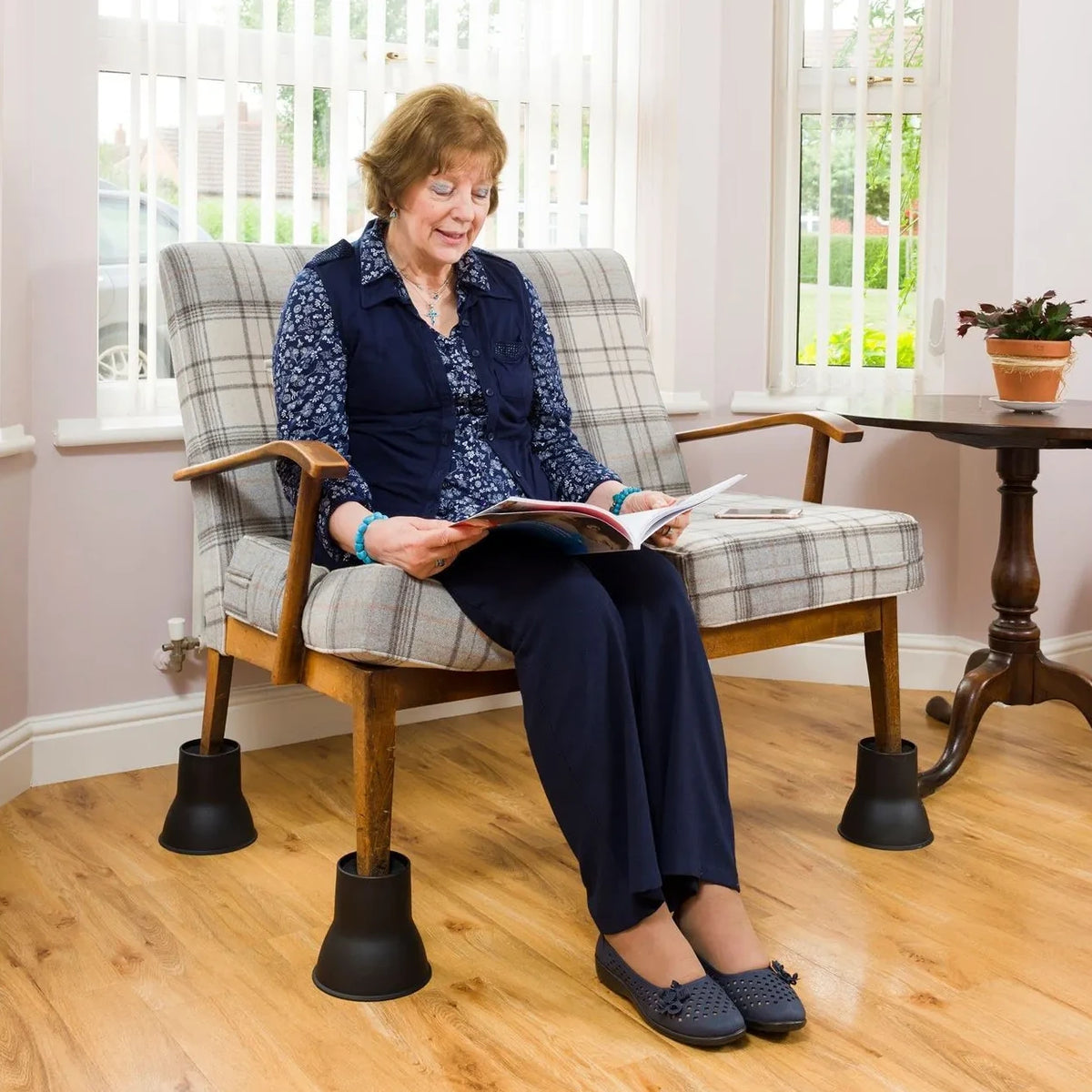 An older woman sits in a wooden armchair with plaid upholstery and Derby Elephant Feet Furniture Raiser 14cm, reading a magazine in a bright living room with wood floors and windows.