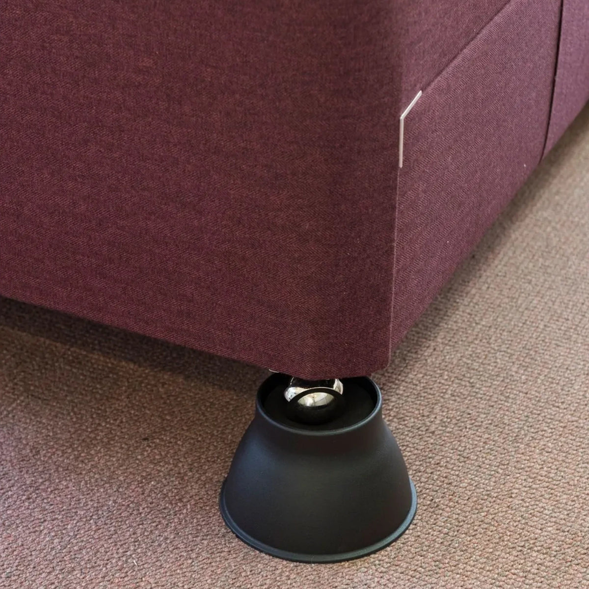 A close-up of a Derby Elephant Feet Furniture Raiser 9cm resting on a black circular floor pad over a textured carpet.