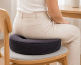 A person seated on a wooden chair on a icare Supporta Donut Cushion charcoal  in a bright indoor room.