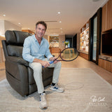 Man sitting on a Theorem Winslow Power Lift Recliner Chair Saddle holding a tennis racket in a modern living room.