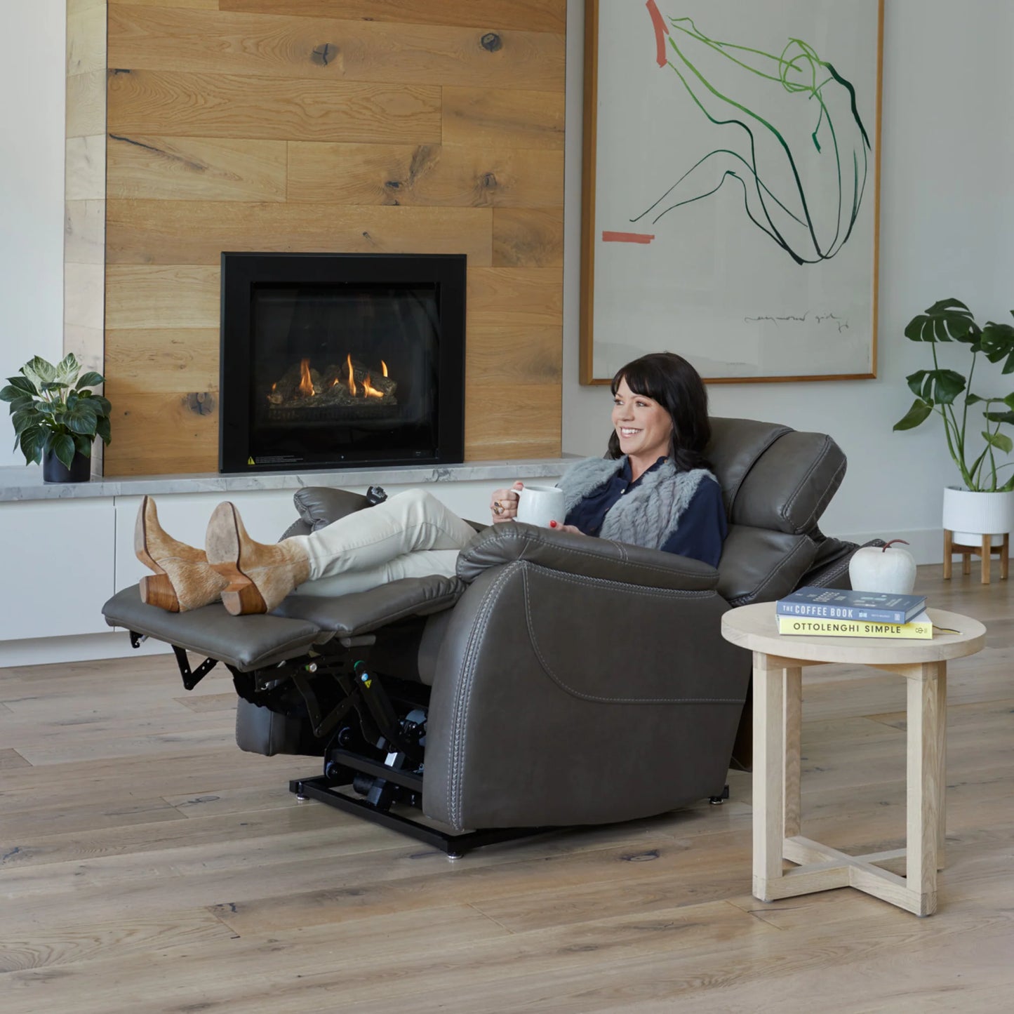 Woman relaxing in a Theorem Winslow Power Lift Recliner Chair in a living room with a fireplace and artwork.