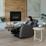 Woman relaxing in a Theorem Winslow Power Lift Recliner Chair in a living room with a fireplace and artwork.