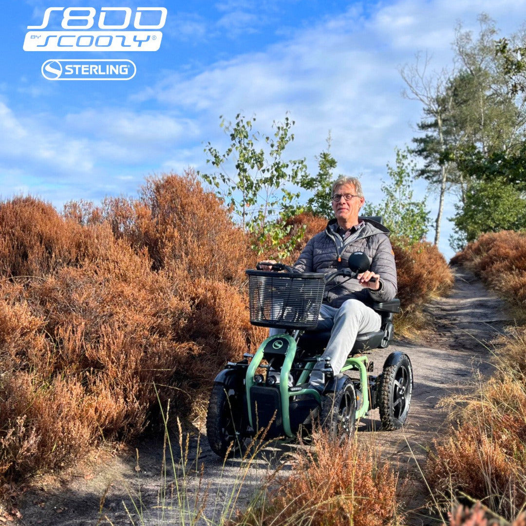 A man rides a green and black STERLING S800 by Scoozy All-Terrain Mobility Scooter on a dirt path surrounded by dry orange-brown heather bushes under a blue sky. Sterling logo is visible on the top left.