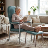 An elderly woman sits in a teal Viking Rehab Chair with a matching Viking Adjustable Legrest in a bright living room, reading a book. The chair has light gray metal legs and armrests, and the room includes a sofa, coffee table, rug, and window.