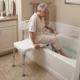 An elderly woman in a beige robe sits on a white Viking Transfer Bench with metal legs, with her feet in the water in a beige tiled bathroom.