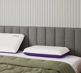 Two white icare Classic Pillows with purple trim, one showing the 'care' logo, arranged on a bed with a gray upholstered headboard and a green quilted blanket.