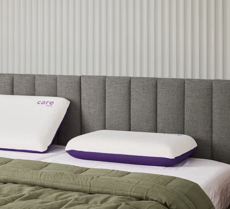 Two white icare Classic Pillows with purple trim, one showing the 'care' logo, arranged on a bed with a gray upholstered headboard and a green quilted blanket.