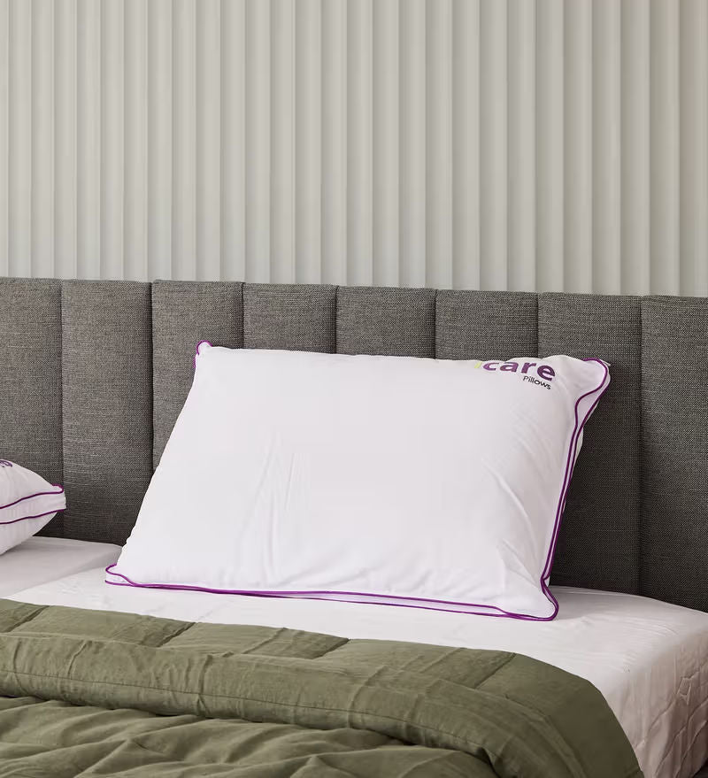 A white icare Cloud Pillow with purple piping and a purple 'icare' logo, resting on a bed with a gray upholstered headboard and an olive green quilt.