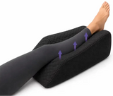 A person's leg and foot resting on a charcoal icare Leg Raiser pillow, with purple arrows indicating leg elevation, on a white background.