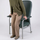 A person wearing an olive green cardigan, tan pants, and brown shoes stands beside a Viking Euro High Back Rehab Chair with a metal frame, holding the armrests against a light gray studio background.