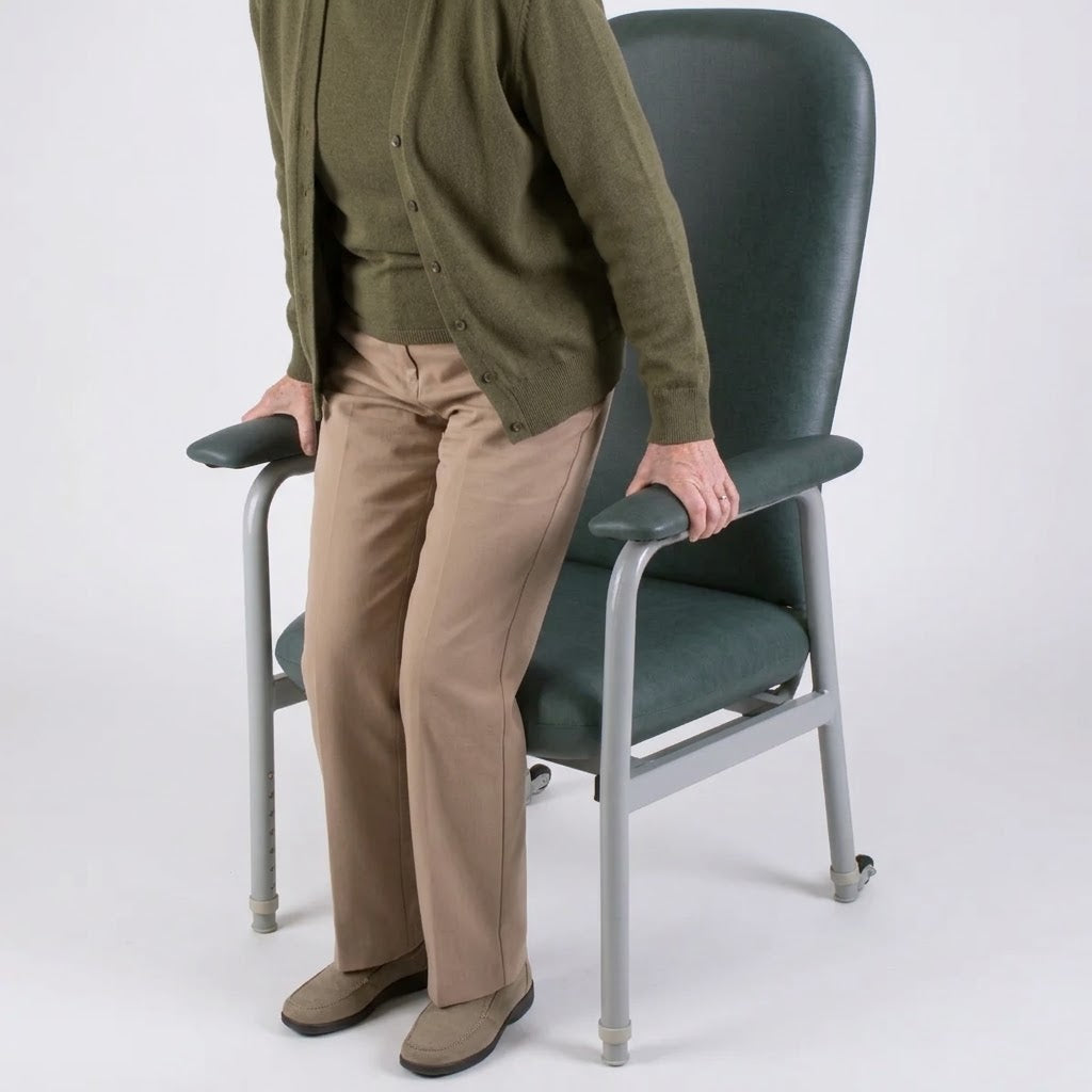 A person wearing an olive green cardigan, tan pants, and brown shoes stands beside a Viking Euro High Back Rehab Chair with a metal frame, holding the armrests against a light gray studio background.
