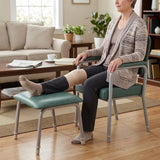 A woman sits in a green Viking Rehab Chair with a matching Viking Adjustable Legrest in a bright living room. Her leg is extended onto the footrest, and she is wearing a beige compression sleeve on her lower leg.