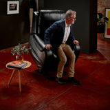 Man standing from a Midnight Theorem Seagrove Power Lift Recliner Chair in a room with red flooring and a small table with books and a plant.