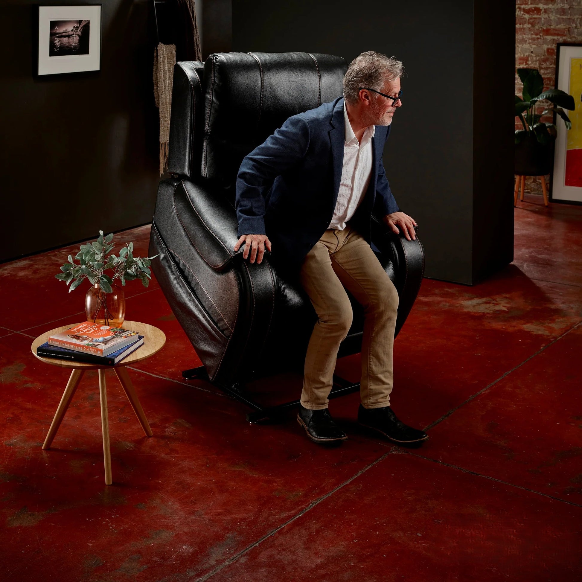 Man standing from a Midnight Theorem Seagrove Power Lift Recliner Chair in a room with red flooring and a small table with books and a plant.
