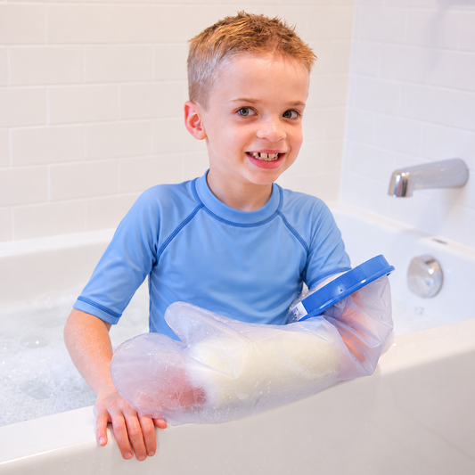 A young boy with short blond hair smiling in a white bathtub, wearing a light blue long-sleeve rash guard with Waterproof Cast Protector on his arm.
