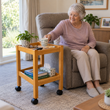 a woman sitting in a recliner with a Tas-Tech Wooden Rehabilitation Utility Trolley without a handle and black wheels in a bright, modern living room. The cart has two shelves, one holding a mug, a small plant, and a tissue box.