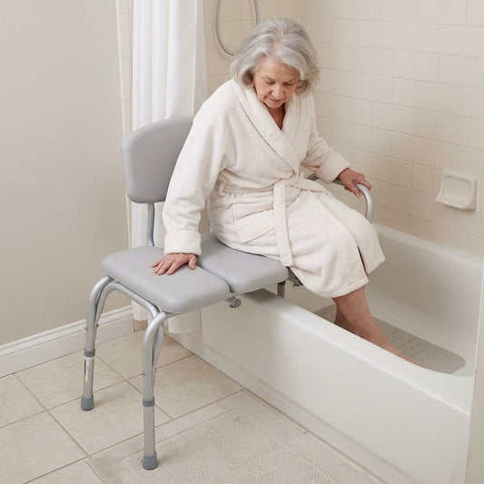 An elderly woman in a cream bathrobe sits on a Chevron Padded Transfer Bench with armrests, with one foot in a white bathtub in a tiled bathroom.