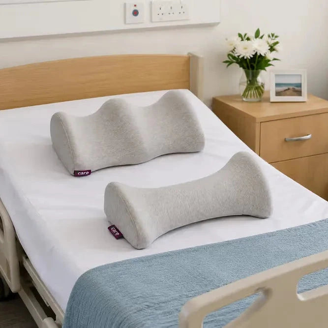 Two gray contoured body pillows- icare Reform Knee Support and icare Reform Lumbar Support placed on a white mattress in a hospital-style bed, with a wooden bedside table, flowers, and a blue chair in the room.