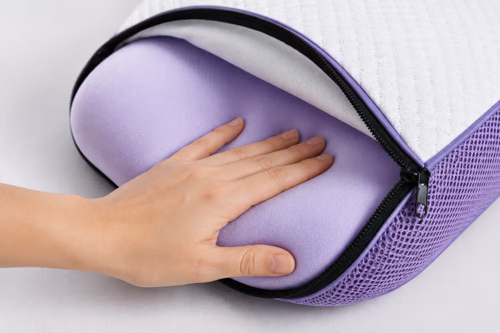 A close-up of a lavender-colored memory foam icare Curve Pillow inner with a black zipper and white top layer, being held by a hand to show its soft texture.