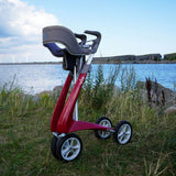 A red byACRE Carbon Ultralight padded backrest for the Wide Track walker positioned on a grassy shoreline with water and a cloudy sky in the background.