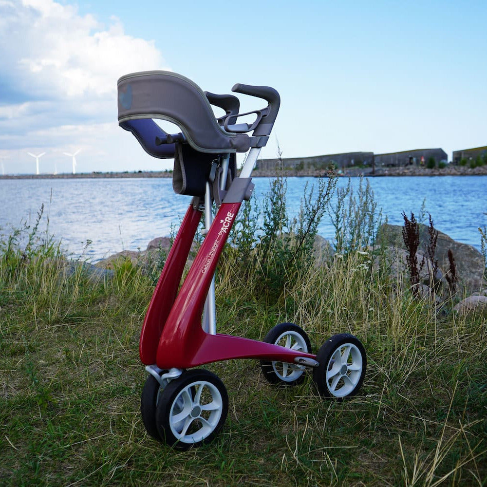 A red byACRE Carbon Ultralight padded backrest for the Wide Track walker positioned on a grassy shoreline with water and a cloudy sky in the background.