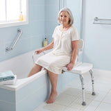 An older woman in a cream nightgown sits on a white Viking Transfer Bench with metal legs in a light blue tiled bathroom.