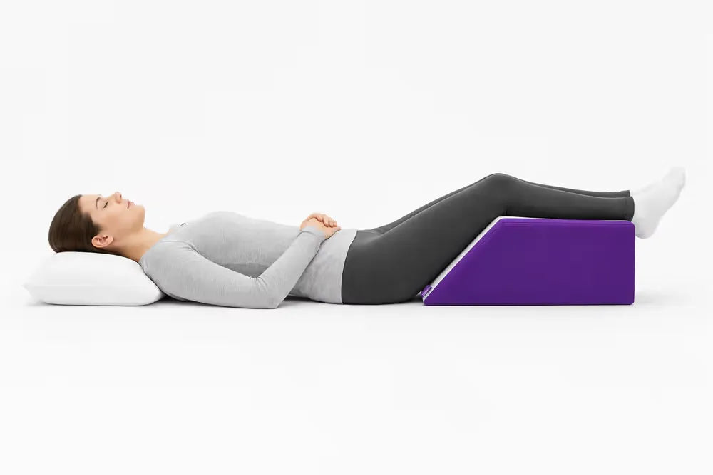 A woman lying on her back with her legs raised on a purple icare Leg Raiser, wearing a gray long-sleeve top, dark leggings, and white socks, set against a plain white studio background.