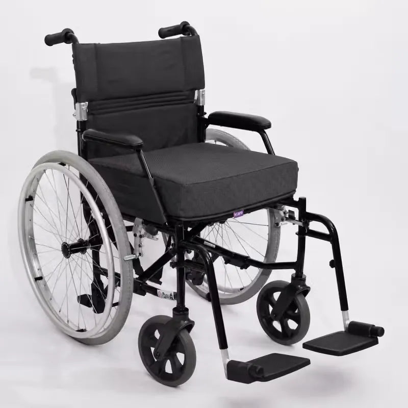 A manual wheelchair with a charcoal icare Supporta Seat Cushion, black armrests, and a black metal frame with silver-spoked rear wheels and black front casters, photographed on a light gray studio background.