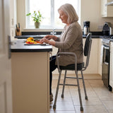 An older woman sits on a Viking Support Stool with Arms while preparing food at a kitchen island. The stool features a padded seat and backrest with a sturdy gray metal frame, set in a bright, modern kitchen.