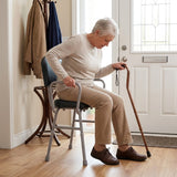 An elderly woman sits on a Viking Support Stool with Arms in a home entryway, holding a wooden cane. She is wearing a beige sweater, beige pants, and brown shoes.