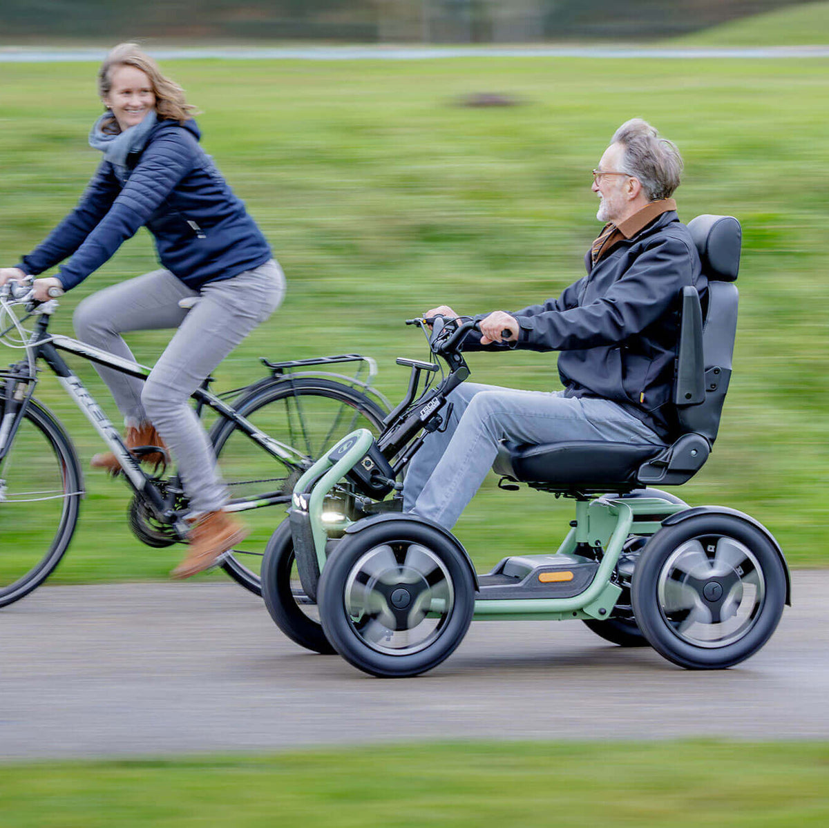 Two people, one on a bicycle and the other in a STERLING S800 by Scoozy All-Terrain Mobility Scooter, riding together on a path.