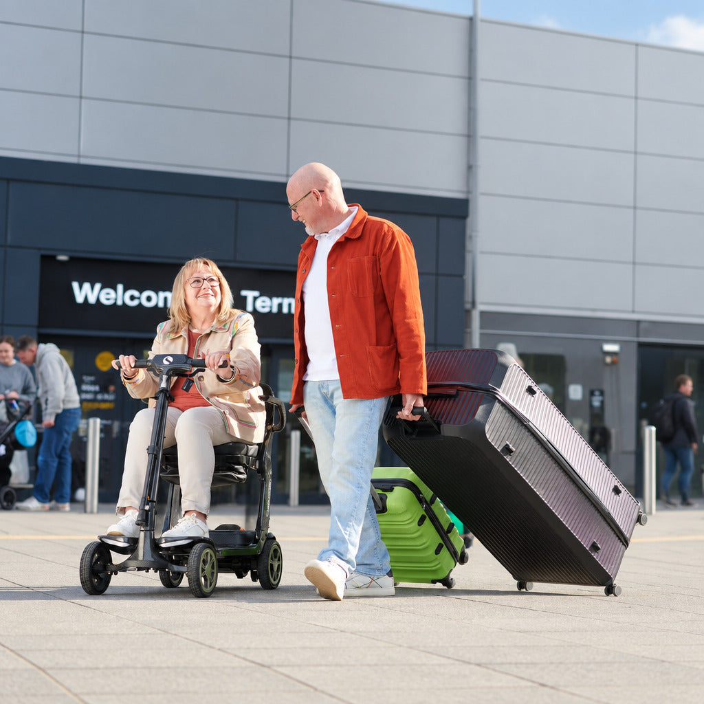 A man in an orange jacket and light blue jeans walks beside a woman using a STERLING S50 Carbon Folding Mobility Scooter, with a large black suitcase in one hand and a green rolling suitcase under the other, in front of a modern building entrance.