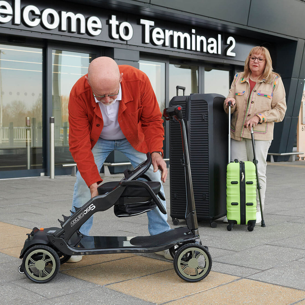 A man in a red jacket folds a STERLING S50 Carbon Folding Mobility Scooter at an airport terminal, with a woman holding a lime green suitcase standing in the background near a 'Welcome to Terminal 2' sign.