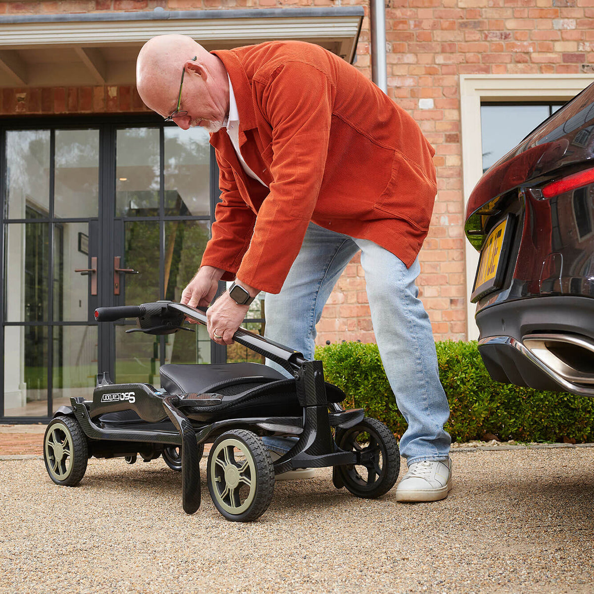 A man in an orange jacket and jeans is folding a STERLING S50 Carbon Folding Mobility Scooter next to a black car in front of a brick house.